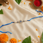 Premium Blue Hamsa Rakhi with Evil Eye charm showcased alongside marigold flowers, diya and traditional Raksha Bandhan décor.
