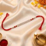 Festive crystal designer Rakhi with silver finish displayed beside diya, kumkum, rice, and marigold flowers