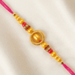 Eshanarya Gold-Tone Oval Designer Rakhi with Pink Thread, Yellow Beads and Red Decorative Accents on Cream Satin Background